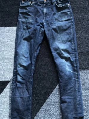 Blå jeans från Nudie Jeans| strl 29/34 - Säljer ett par klassiska blå jeans från Nudie Jeans med snygg tvätt och raka ben. Jeansen har fem fickor, läderpatch bak i midjan och kontrastsömmar på bakfickorna. Perfekta till en avslappnad stil och tillverkade i slitstarkt bomullsmaterial. Dom sitter slim på