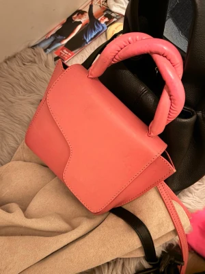 Atp ateiler - Säljer en superfin rosa axelväska med unikt twistat handtag och asymmetrisk lock. Väskan har en clean look och är gjord i slätt syntetmaterial. Perfekt storlek för det viktigaste och har en lång axelrem så du kan bära den crossbody.