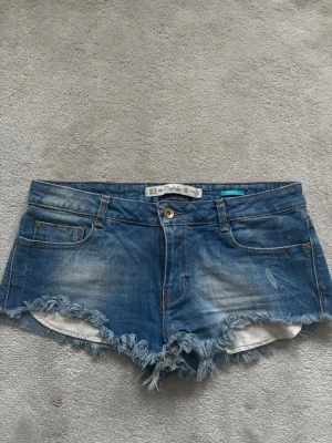 Blå jeansshorts från Zara Trafaluc med fransar - Säljer ett par blå jeansshorts från Zara Trafaluc med fransiga kanter och klassisk femficksdesign. Shortsen har låg midja, dragkedja och knapp framtill samt slitna detaljer. Köpta på Vinted.
