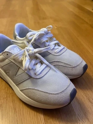 Vita New Balance 370 sneakers med mockadetaljer - Säljer ett par stilrena New Balance sneakers i vitt med ljusgrå detaljer. Skorna har ovandel i mesh och mocka, klassisk snörning och rund tå. Tjock sula för extra komfort och logga på både plös och häl. Perfekta för dig som gillar en clean och sportig look.