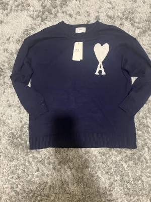 Mörkblå stickad tröja från Ami - Säljer en mörkblå stickad tröja från Ami med ikoniskt vitt hjärta och A på bröstet. Tröjan har rund hals, ribbade muddar och lång ärm. Perfekt för dig som vill ha en clean men ändå statement look.