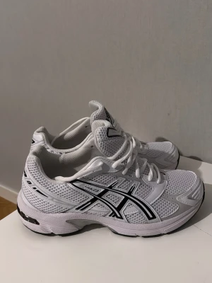 Vita Asics sneakers - Säljer ett par fräscha vita Asics sneakers med svart logga på sidan. Skorna har ventilerande mesh-ovandel, klassisk snörning och en chunky sula för extra komfort. Perfekta för dig som gillar sportig stil och vill ha något bekvämt till vardags.