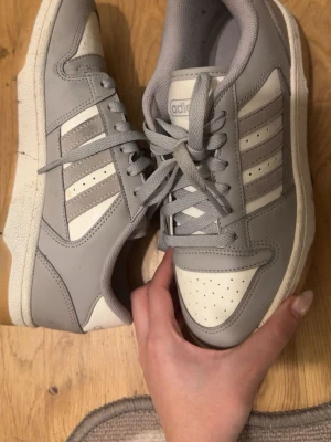 Gråvita Adidas sneakers med snörning - Säljer ett par stilrena Adidas sneakers i grått och vitt. Skorna har klassiska tre ränder på sidan, perforerad tå och tjock vit sula. Ovandelen är i syntetmaterial och mockadetaljer, med skön vadderad insida och snörning framtill. Perfekta till vardagslooken!