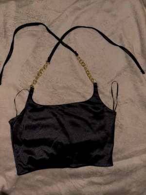 Svart satin linnetopp med guldkedjor - Säljer en svart linnetopp i glansigt satinmaterial med tunna axelband och snygga guldfärgade kedjedetaljer framtill. Toppen är croppad och har en elegant look som sticker ut. Perfekt för dig som vill ha något extra till outfiten.