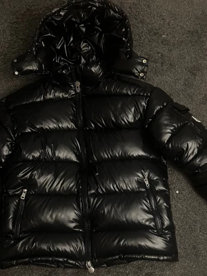 Svart Moncler pufferjacka med huva - Säljer en svart pufferjacka från Moncler med glansig finish och huva. Jackan har dragkedja framtill, två sidofickor med dragkedja och en ficka på ärmen. Insidan har Monclers klassiska serietidningsmärke. Perfekt för kalla dagar.