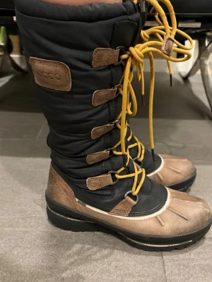 Svarta vinterstövlar från ECCO - Säljer ett par höga vinterstövlar från ECCO i svart och brunt med gula snören. Utsidan är i slitstark textil och skinn, och insidan är fodrad med mjuk ull för extra värme. Stövlarna har grov sula och snörning hela vägen upp för bästa passform. 