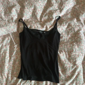 Svart spetslinnetopp  - Svart linne från The Black Label Collection (lager 157) med smala axelband och v-ringning dekorerad med spets. Toppens material känns mjukt och stretchigt. Kontakta mig om du har några frågor ❤️❤️
