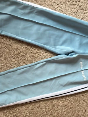 Ljusblåa Palm Angels mjukisbyxor - Säljer ett par ljusblåa mjukisbyxor från Palm Angels med vita och svarta ränder längs sidorna. Byxorna har resår i midjan, raka ben och sidofickor. Palm Angels-loggan är broderad i vitt på ena benet. Perfekta för en chill och sportig look.