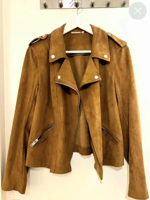 Brun mocka bikerjacka från KappAhl - Snygg bikerjacka i brun mocka-look från KappAhl. Jackan har bred krage, axelklaffar och silverfärgade dragkedjor på fickorna. Perfekt för dig som vill ha en cool och tidlös stil. Långärmad och med klassisk bikerdesign.