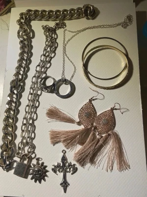Smyckespaket med kedjor och örhängen - Flera smycken säljes tillsammans: två grova silverfärgade kedjor, halsband med berlock, en ring, två guldiga armband och ett par stora örhängen med beige tofsar och metallmönster. Inkluderar även sol- och korsberlocker. Perfekt för dig som gillar att mixa stilar.