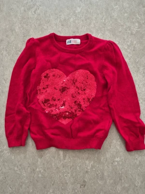 Stickad tröja - Supersöt röd stickad tröja från H&M med ett stort hjärta i röda paljetter på bröstet. Tröjan har rund halsringning och långa ärmar med lätt puff vid axlarna. Perfekt för dig som vill ha något gulligt och färgglatt i garderoben!