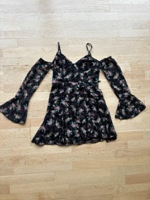 Svart blommig kortklänning med snörning - Supersöt svart kortklänning med blommigt mönster . Klänningen har offshoulder långa ärmar med volangkant, smala axelband och snörning i ryggen. En ögla saknas för snörningen på höger sida, se bild 4. 