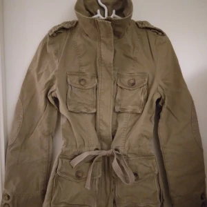 Beige parkas / sommarjacka / vårjacka / cargojacka / utilityjacka. Strlk: XS. Felfri. - Beige parkas / sommarjacka / vårjacka / cargojacka / utilityjacka. Strlk: XS.  Fint skick, knappt använd, felfri. Kraftigare bomullstyg samt fint äppelmönstrat foder inuti. Flera fickor. Stängs med dragkedja och har knytband i midjan. FRAKT TILLKOMMER.