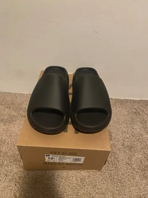 Adidas Yeezy Slide Onyx strl 44.5 - Säljer ett par Adidas Yeezy Slide i färgen Onyx. Slidesen har en clean, minimalistisk look med öppen tå och räfflad sula för extra grepp. Tillverkade i syntetmaterial och perfekta för chill dagar. Kommer i originalkartong.