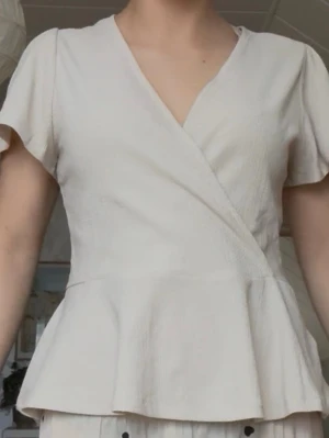 Krämvit omlottopp med volang - Säljer en krämvit omlottopp med korta ärmar och volang nedtill från Holly & Whyte. Toppen har v-ringning och en mjuk, lätt struktur i tyget. Perfekt till våren och sommaren 💕💕