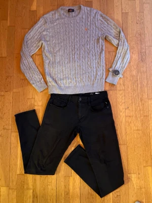 Grå kabelstickad tröja Morris - ‼️Tröjan är XL men passar även LSäljer en grå kabelstickad tröja från Morris i storlek XL. Tröjan har rund hals, ribbade muddar och en orange logga broderad på bröstet. Perfekt till jeans eller chinos för en clean look. Modellen är 182