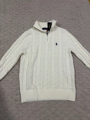 Vit kabelstickad tröja Polo Ralph Lauren - Kabelstickad vit tröja från Polo Ralph Lauren med halv dragkedja och klassisk broderad logga på bröstet. Tröjan har hög krage, ribbade muddar och är långärmad. Perfekt för dig som gillar en clean och tidlös look.