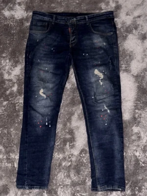 Mörkblå Dsquared2 jeans med färgstänk storlek 52 - Säljer ett par mörkblå jeans från Dsquared2 med coola färgstänk i vitt och rött på framsidan. Jeansen har klassisk femficksdesign, rakt snitt och detaljerad söm. Märkespatch i rött bak och Dsquared2-tag vid bakfickan. Perfekta för dig som gillar streetwear vibes.