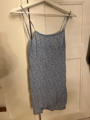 Blommig blå kortklänning från SHEIN - Supersöt blå kortklänning från SHEIN i storlek XS. Klänningen har tunna axelband, blommigt mönster i vitt och gult samt dragkedja i sidan. Perfekt för sommarens alla dagar och ger en fräsch och ungdomlig vibe.