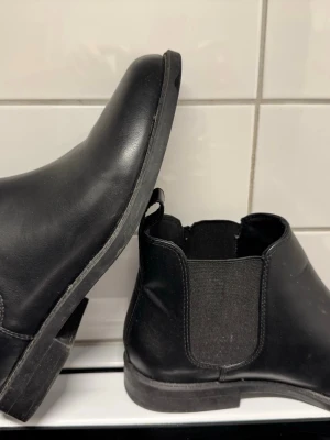 Svarta chelsea boots från Divided - Säljer ett par klassiska svarta chelsea boots från Divided. Skorna har elastiska paneler på sidorna, rund tå och en låg klack. Tillverkade i syntetmaterial med slät finish och dragflik bak för enkel påtagning. Perfekta till jeans eller kjol.