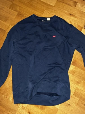 Mörkblå sweatshirt från Levi's - Klassisk mörkblå sweatshirt från Levi's i bomull med rund hals och diskret röd Levi's-logga på bröstet. Tröjan har långa ärmar och ribbade muddar vid ärmslut och nederkant. Perfekt för en avslappnad och stilren look.