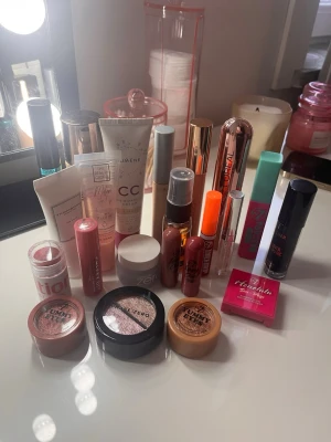 Stort sminkpaket med flera märken - Säljer ett stort sminkpaket med blandade produkter: foundation, CC-cream, primer, mascara, läppstift, läppglans, rouge, bronzer, highlighter och ögonskuggor. Färgerna går i nude, rosa, brons och guld. Inkluderar både flytande och puderprodukter från populära märken.