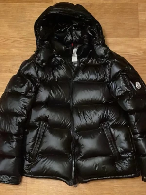 Moncler Maya dunjacka (S:3) - Säljer en svart pufferjacka från Moncler med stor vadderad huva och två snedställda fickor med dragkedja. Jackan har en glansig finish och klassisk Moncler-logga på ärmen. Perfekt för kalla dagar och riktigt snygg till streetwear. Jackan är i nyskick och är i size 3. Modellen på bilden är 180cm lång. Pris kan diskuteras.