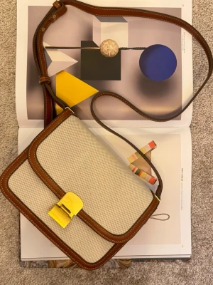 Mango axelväska - Snygg beige axelväska med bruna skinnkanter och justerbar axelrem. Väskan har en guldfärgad metallspänne framtill och är tillverkad i en kombination av textil och skinn. Klassisk och stilren design som passar till många olika looks.