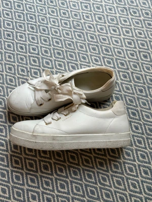 Vita sneakers från Gant med breda snören - Säljer ett par klassiska vita sneakers från Gant med breda, platta snören och guldfärgade detaljer vid snörningen. Skorna har en rund tå och platt sula, tillverkade i skinn för en clean och stilren look.