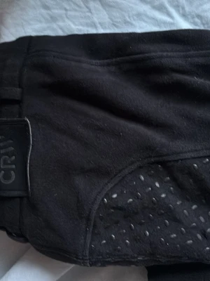Svarta ridbyxor från CRW - Säljer ett par svarta ridbyxor från CRW med helskoning i silikon för extra grepp. Byxorna har dragkedjeficka framtill och bälteshällor i midjan. Snygg och funktionell design som passar perfekt till ridning.