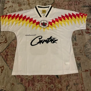 Cortez vit fotbollströja med grafiskt mönster - Vit fotbollströja från Cortez med coolt grafiskt mönster i rött och gult över axlarna. Broderad logga med stjärna och svart tryck på bröstet. Svartvit krage och lätt, ventilerande polyester. Perfekt för dig som gillar retrostil och sport.