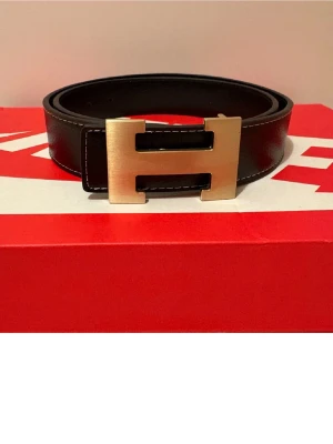 Svart  skärp med H-spänne  - Svart skärp från h&m med ikoniskt H-spänne i guldtonad metall. Skärpet är tillverkat i äkta skinn och har vita sömmar längs kanterna för en snygg kontrast. Perfekt accessoar för att lyfta din outfit med lyxig känsla.