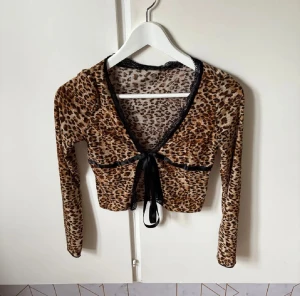 Leopardmönstrad croppad bolerotopp - Säljer en croppad bolerotopp med leopardmönster i bruna och beige toner. Toppen har lång ärm, v-ringning med svart spetskant och ett svart satinband som knyts framtill. Materialet är mjukt och stretchigt, perfekt för att ge en cool vibe till din outfit.