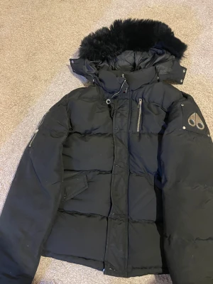 Svart Moose Knuckles dunjacka med päls - Svart dunjacka från Moose Knuckles med fluffig svart päls på huvan, coola metalldetaljer på ärmen och dragkedjor vid ärmslut. Jackan har flera fickor med dragkedja och en broderad hockeypatch på insidan. Perfekt för kalla dagar och riktigt snygg streetstil.