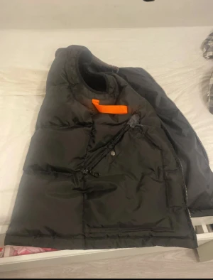 Svart dunväst från Parajumpers - Svart dunväst från Parajumpers i slim fit med orange detalj vid kragen. Västen har dragkedja och knappstängning på fickorna, quiltad design och mjukt foder. Perfekt för kalla dagar när du vill ha en cool och funktionell look.