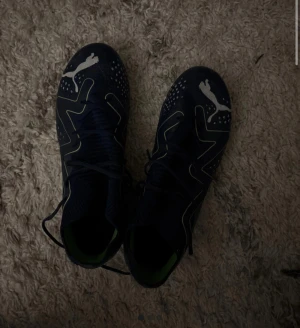 Mörkblå fotbollsskor från Puma strl 38 - Säljer ett par mörkblå fotbollsskor från Puma i storlek 38. Skorna har vita Puma-loggor, snörning och detaljer i limegrönt på sulan och insidan. Snyggt mönster med prickar och linjer på ovansidan. Använda en gång men då dem inte passar aldrig använda igen.