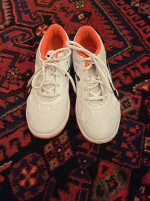 Vita och orangea gympaskor från Puma - Säljer ett par fräscha gympaskor från Puma i vitt med svart logga och orange insida samt sula. Skorna har snörning och en cool struktur på ovansidan. Perfekt för dig som vill sticka ut på planen med snygg design och bra grepp.