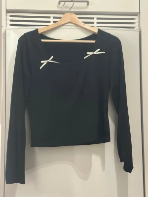 Svart långärmad topp med rosetter - Svart långärmad topp med två vita rosetter vid den fyrkantiga halsringningen. Modellen är croppad och har en stilren, enkel design som passar perfekt till jeans eller kjol. Materialet känns mjukt och stretchigt, vilket gör den bekväm att bära hela dagen.