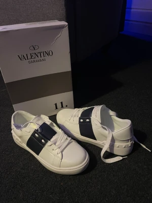 Valentino Garavani vita sneakers - Säljer ett par fräscha Valentino Garavani sneakers i vitt skinn med breda mörkblå band och coola nitar på sidan. Klassisk rund tå, platt sula och snörning framtill. Märkeslogga på plösen och insidan. Perfekt för dig som gillar lyxiga detaljer och stilren design.