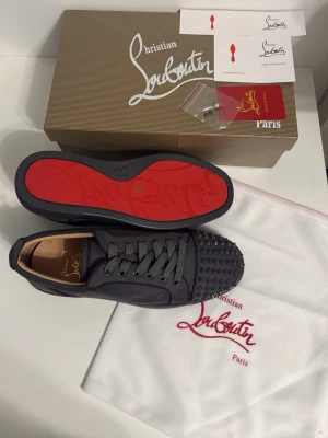 Gråa sneakers från Christian Louboutin med spikar - Säljer ett par gråa sneakers från Christian Louboutin med ikonisk röd sula och coola nitar på tån. Skorna har snörning och är tillverkade i skinn med en platt sula. Perfekt för dig som vill sticka ut med lyxig streetstyle.