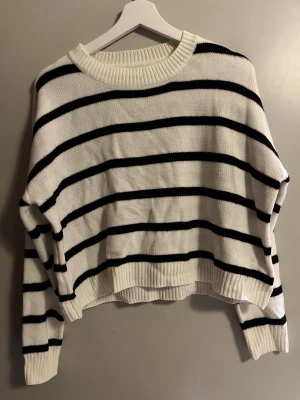 Vit och svart randig stickad tröja - Snygg stickad tröja i vit med svarta horisontella ränder. Modellen är loose fit med lång ärm och ribbad rund hals samt muddar vid ärmslut och nederkant. Perfekt för dig som gillar en chill och trendig stil.