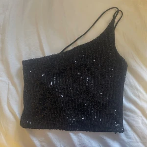 Svart paljett topp med oneshoulder - Säljer en svart glittrig topp med paljetter och oneshoulder-design. Toppen har smala axelband och är croppad, perfekt för att sticka ut på festen. Den glittriga ytan ger ett lyxigt intryck och passar dig som gillar att synas.