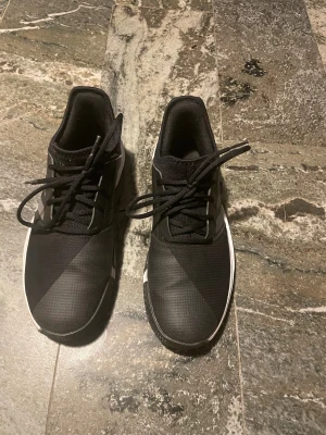 Svarta Adidas sneakers med snörning - Säljer ett par svarta Adidas sneakers med klassiska tre ränder på sidan. Skorna har rund tå, platt sula och snörning. Ovandelen är i syntetmaterial och mesh för bra andning. Sulan är vit med mönstrad undersida för extra grepp. Perfekt för träning eller chill. Anpassade för inomhus aktiviteter, men kan användas utomhus som man väljer det. Herr storlek 41, Dam storlek 39.