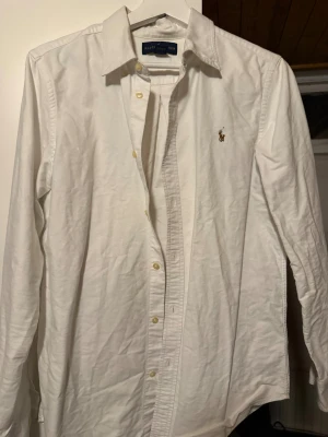 Vit skjorta från Ralph Lauren - Klassisk vit skjorta från Ralph Lauren med broderad logga på bröstet. Skjortan har lång ärm, knappar framtill och en snygg krage. Tillverkad i mjuk bomull och har en clean, tidlös look som funkar till allt.