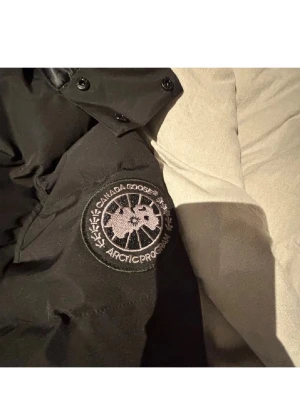 Svart pufferjacka från Canada Goose - Svart pufferjacka från Canada Goose med ikonisk patch på ärmen. Jackan har huva, dragkedja och knappar framtill. Fodret är glansigt och jackan är rejält vadderad för extra värme. Perfekt för kalla dagar och har en clean, stilren look.