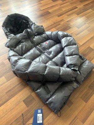 Grå dunjacka från Polo Ralph Lauren - Snygg grå dunjacka från Polo Ralph Lauren med glansig finish, huva och dragkedjor. Jackan har det klassiska röda Polo-logot broderat på bröstet och justerbara muddar. Perfekt för kalla dagar och riktigt stilren look.