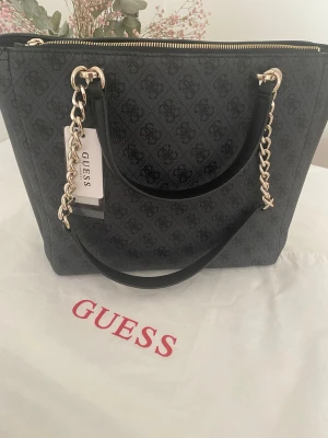 Svart axelväska från Guess med kedja - Snygg svart axelväska från Guess med mönstrad yta och guldfärgade kedjedetaljer. Väskan har dragkedja upptill, flera fack inuti och ett stilrent Guess-emblem framtill. Materialet är syntet med en lyxig känsla och väskan har en klassisk, fyrkantig form.