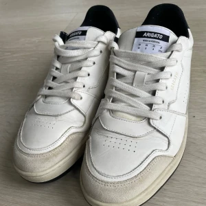 Vita sneakers från Axel Arigato i storlek 42. Pris kan ändras via prisförslag.  - Snygga vita sneakers från Axel Arigato med svart häl och beige detaljer på tån. Skorna har snörning, perforerade partier på sidorna och platt sula i svart och beige. Tillverkade i skinn och mocka, low cut modell med stilren design. Lägg prisförslag om ni vill. 