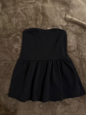 Svart axelbandslös kortklänning - Snygg svart axelbandslös top med voluminös kjol. Modellen är kort och har en stilren, enkel design 