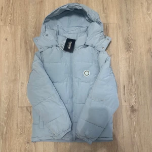 Ljusblå pufferjacka från Trapstar - Trapstar pufferjacka i ljusblå färg med stor huva och dragkedja framtill. Jackan har broderad Trapstar-logga i svart på ryggen och rund patch på bröstet. Tillverkad i syntetmaterial, quiltad design och elastiska muddar vid ärmslut. Perfekt för kalla dagar.